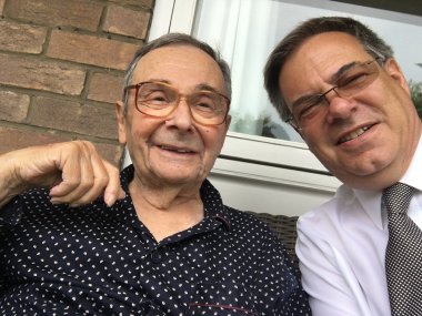 Ein älterer Mann mit gemustertem Hemd und Brille sitzt lächelnd neben einem Mann im weißen Hemd und Krawatte, der ein Selfie auf einer Terrasse aufnimmt.