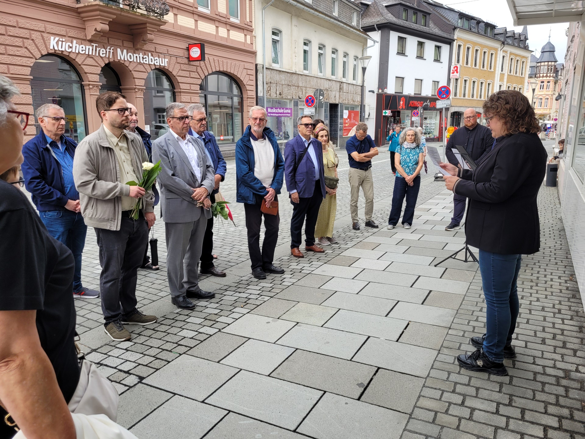 Eine Gruppe von Menschen steht aufmerksam auf einem Gehweg in einer deutschen Innenstadt, während eine Frau mit Mikrofon und Notizen vor ihnen spricht.