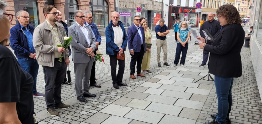Eine Gruppe von Menschen steht aufmerksam auf einem Gehweg in einer deutschen Innenstadt, während eine Frau mit Mikrofon und Notizen vor ihnen spricht.