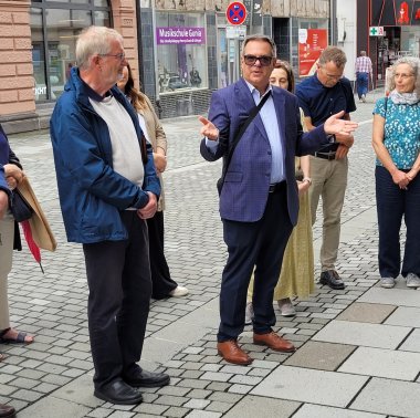 Ein Mann in blauem Sakko und Sonnenbrille spricht gestikulierend vor einer Gruppe von Zuhörern auf einem Gehweg, während die anderen aufmerksam zuhören.