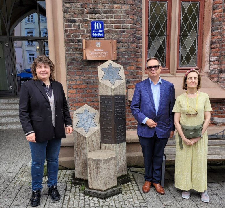 Drei Personen stehen vor einer Gedenkstele mit eingravierten Davidsternen und Namen, an einer roten Backsteinmauer mit gotischen Fenstern und einem blauen Straßenschild „10, Großer Markt“.