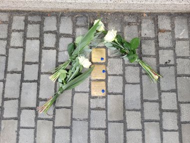 Drei Stolpersteine mit gravierten Namen und Lebensdaten sind in das Pflaster eingelassen, daneben liegen drei weiße Rosensträuße als Zeichen des Gedenkens.