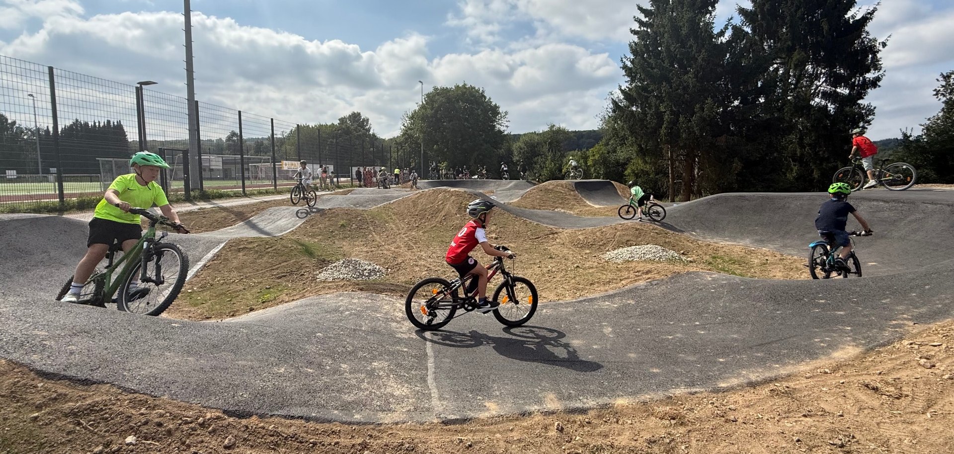 Kinder fahren mit Fahrrädern über einen asphaltierten Pumptrack mit Hügeln und Kurven bei sonnigem Wetter. Im Hintergrund sind weitere Fahrerinnen und Fahrer sowie ein Sportplatz und Bäume zu sehen.