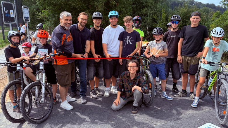 Gruppe von Jugendlichen und Erwachsenen mit Fahrrädern bei der feierlichen Eröffnung eines Bikeparks, während zwei Männer ein rotes Band durchschneiden. Alle tragen Helme und lächeln in die Kamera.