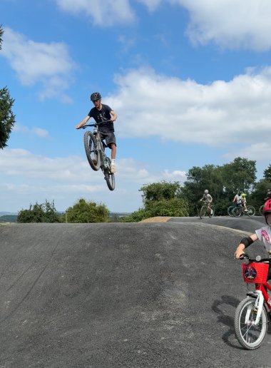 Ein Jugendlicher springt mit seinem Mountainbike spektakulär über eine Erhebung auf dem Pumptrack. Weitere Kinder und Jugendliche fahren im Hintergrund, während ein kleines Kind mit rotem Fahrrad und Helm rechts im Bild zu sehen ist.
