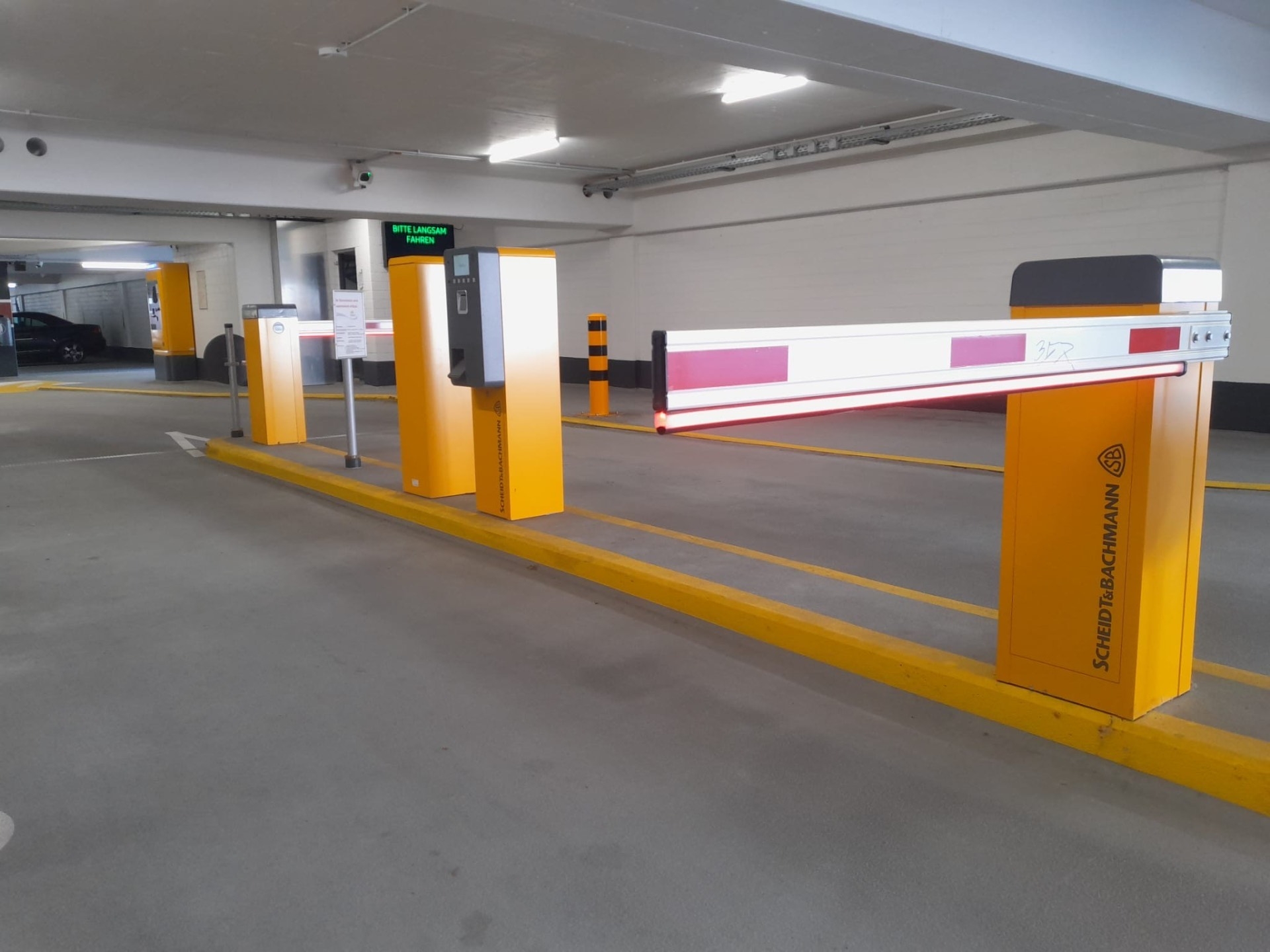 Ein Parkhaus mit einer geschlossenen Schranke von Scheidt & Bachmann, flankiert von gelben Säulen mit Ticketautomat und Display. Auf dem Display steht in Grün "BITTE LANGSAM FAHREN", während rote LED-Streifen die Schranke beleuchten.