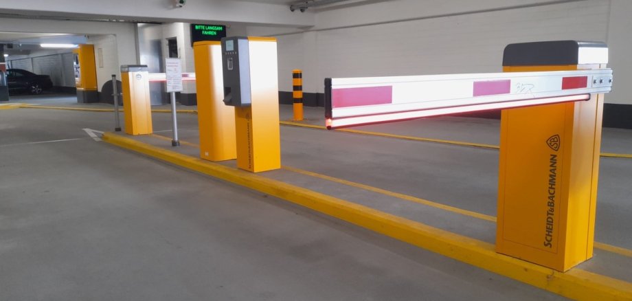 Ein Parkhaus mit einer geschlossenen Schranke von Scheidt & Bachmann, flankiert von gelben Säulen mit Ticketautomat und Display. Auf dem Display steht in Grün "BITTE LANGSAM FAHREN", während rote LED-Streifen die Schranke beleuchten.