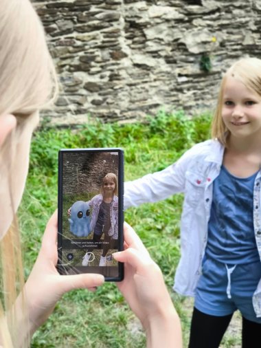 Ein Mädchen wird mit dem Smartphone fotografiert, auf dem Display erscheint neben ihr eine blaue, cartoonartige Augmented-Reality-Figur mit großen Augen. Im Hintergrund ist eine alte Steinmauer und grüne Wiese zu sehen.