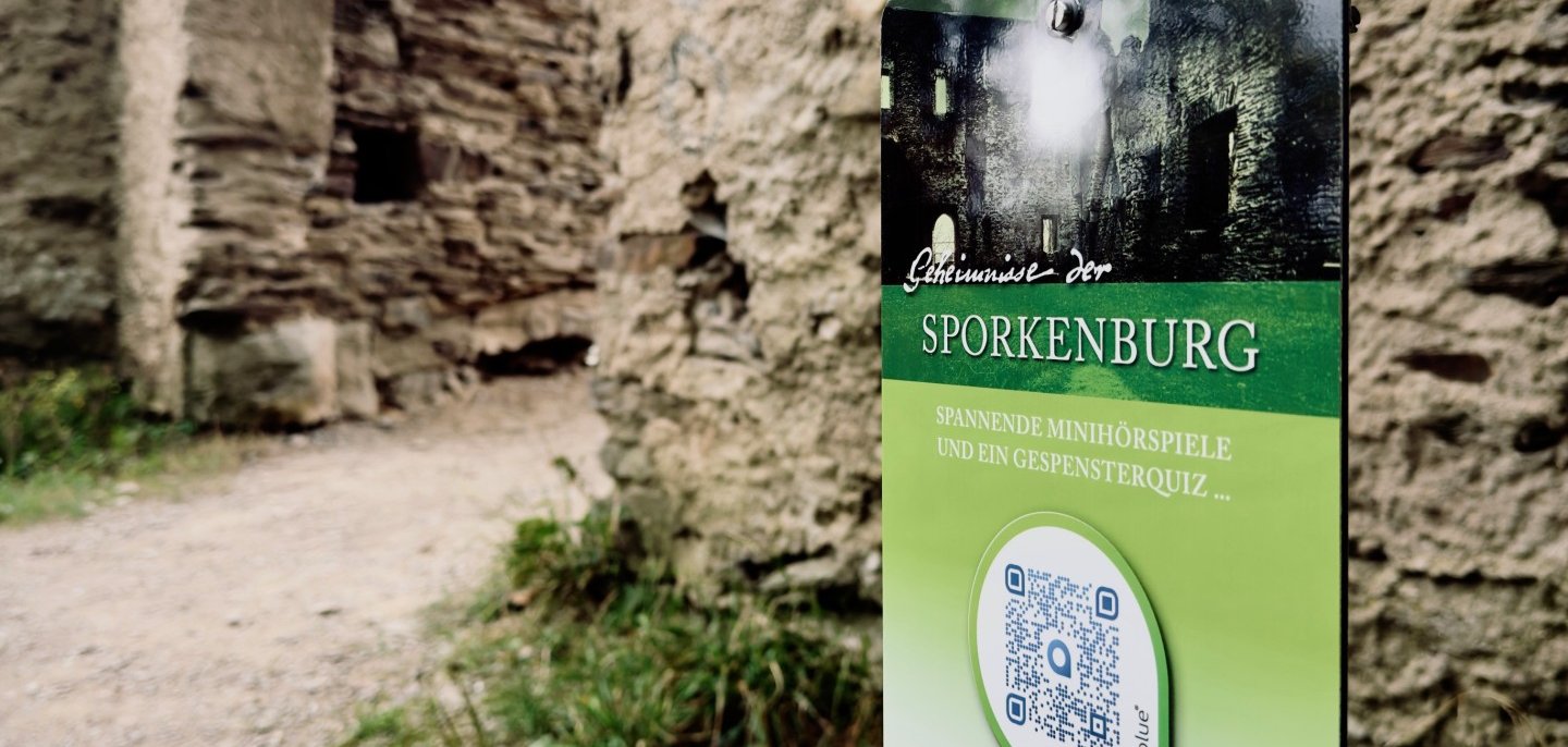Nahaufnahme eines grünen Infoschildes mit der Aufschrift „Sporkenburg – Spannende Minihörspiele und ein Gespensterquiz“ vor dem Eingang der Burgruine. Darunter ist ein großer QR-Code zum Scannen angebracht.