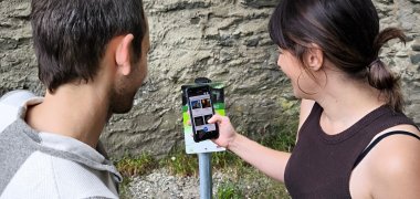 Zwei Personen scannen mit einem Smartphone einen QR-Code auf einem Schild vor einer alten Steinmauer. Das Display zeigt eine Auswahl an Inhalten, die vermutlich zur Sporkenburg gehören.