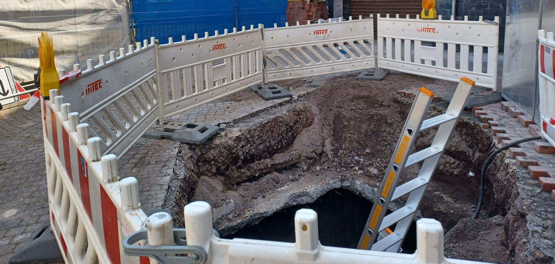 ine abgesperrte Baustelle in der Innenstadt mit aufgerissenem Boden und einer Baugrube, in der eine Leiter steht. Um die Baugrube sind weiße Absperrgitter mit Warnleuchten aufgestellt.