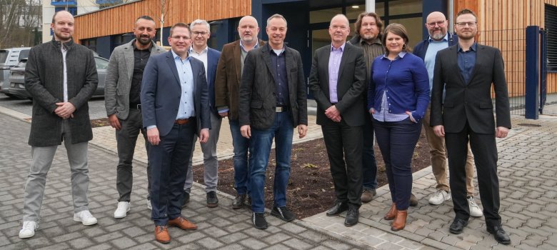 Eine Gruppe von elf Personen steht vor einem modernen Holz- und Glasgebäude für ein Gruppenfoto. Die Personen tragen überwiegend Business-Kleidung, darunter Anzüge, Blazer und Hemden, und schauen freundlich in die Kamera. Im Hintergrund sind ein Parkplatz mit Autos und ein mehrstöckiges Gebäude mit blauen Fenstern und roten Dächern zu sehen.