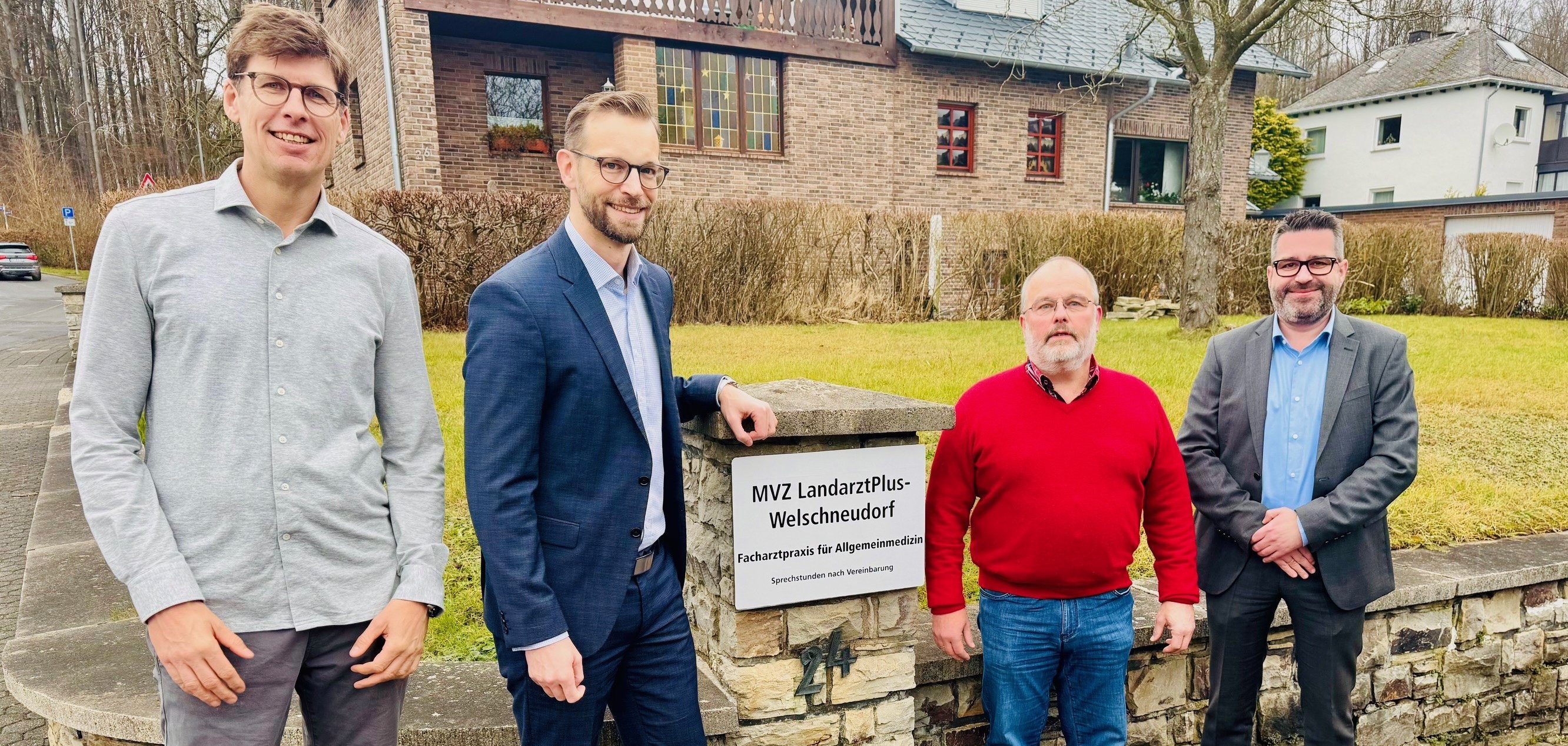 Vier Männer stehen vor einem Gebäude neben einem Schild mit der Aufschrift 'MVZ LandarztPlus-Welschneudorf Facharztpraxis für Allgemeinmedizin.' Die Szene spielt sich auf einem gepflasterten Weg vor einer Steinmauer und einer Rasenfläche ab, mit Wohnhäusern im Hintergrund.