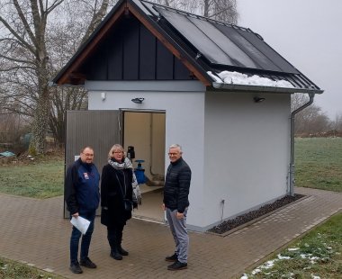 Eine kleine graue Pumpstation mit schrägem Dach und Solarpanelen auf der Oberseite, umgeben von einer winterlichen Landschaft mit kahlen Bäumen. Vor der Station stehen drei Personen, zwei Männer und eine Frau, die lächeln und Papiere in der Hand halten. Das Bild zeigt eine technische Einrichtung in einer ländlichen Umgebung.