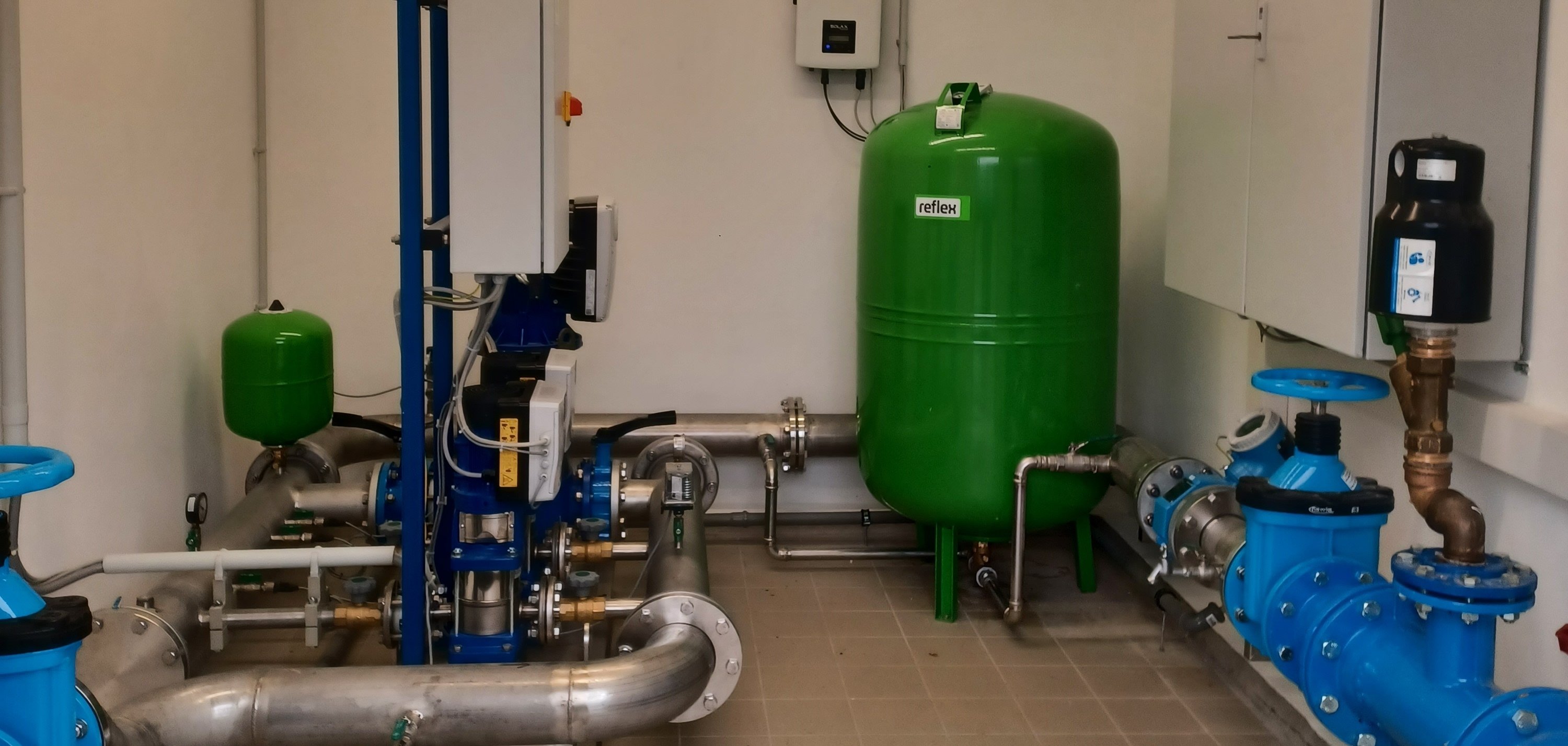 Der Innenraum einer Pumpstation mit moderner technischer Ausstattung. Sichtbar sind blaue und grüne Druckbehälter, silberne Rohrleitungen und Steuergeräte an den Wänden. Der Raum wirkt sauber und gut organisiert, mit klar strukturierten Anlagen zur Wasser- oder Druckregelung.