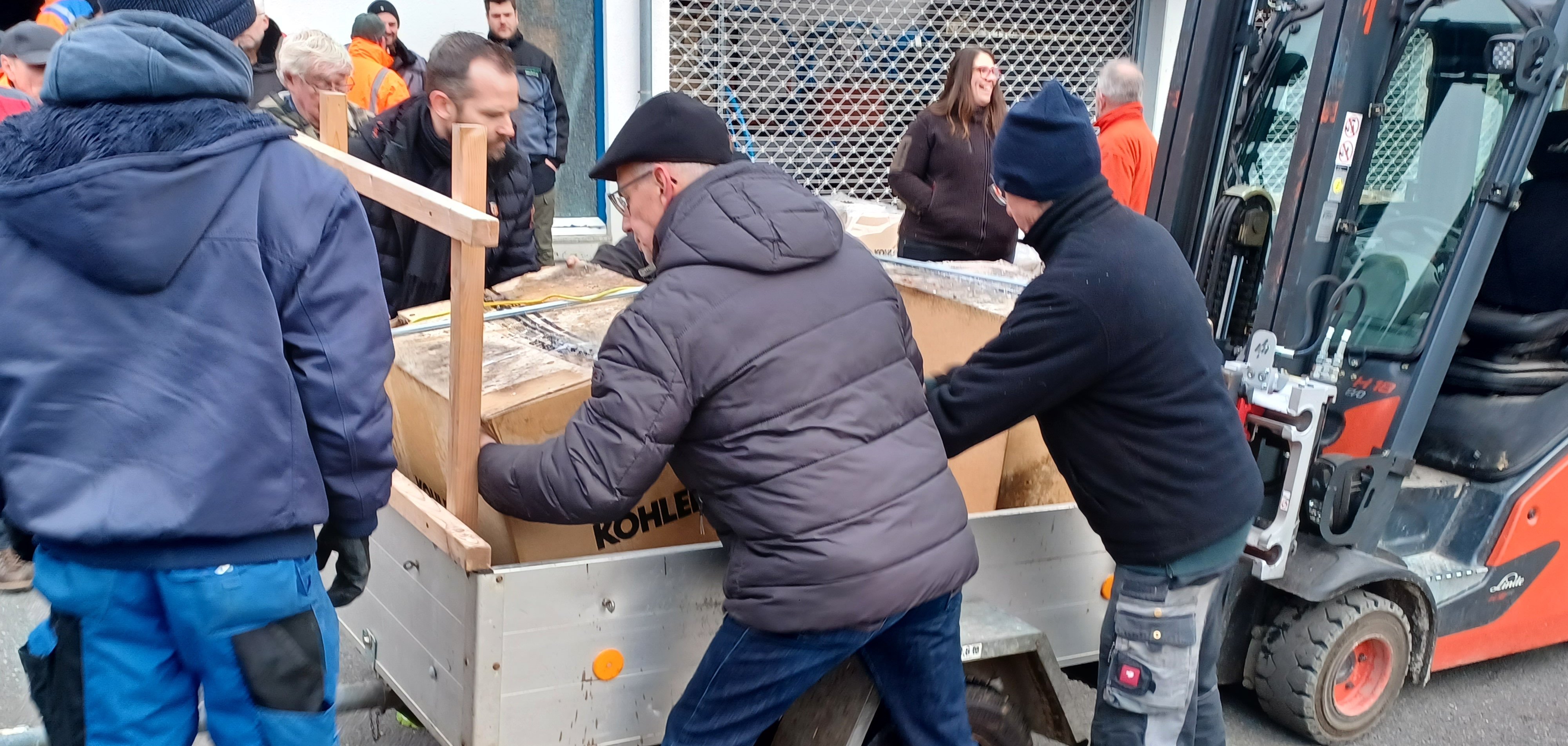 Mehrere Männer laden eine große Holzkiste aus einem Anhänger. Sie tragen warme Arbeitskleidung, während im Hintergrund ein Gabelstapler und mehrere Personen zu sehen sind. Die Szene spielt sich vor einer Halle mit einem geschlossenen Rolltor ab.