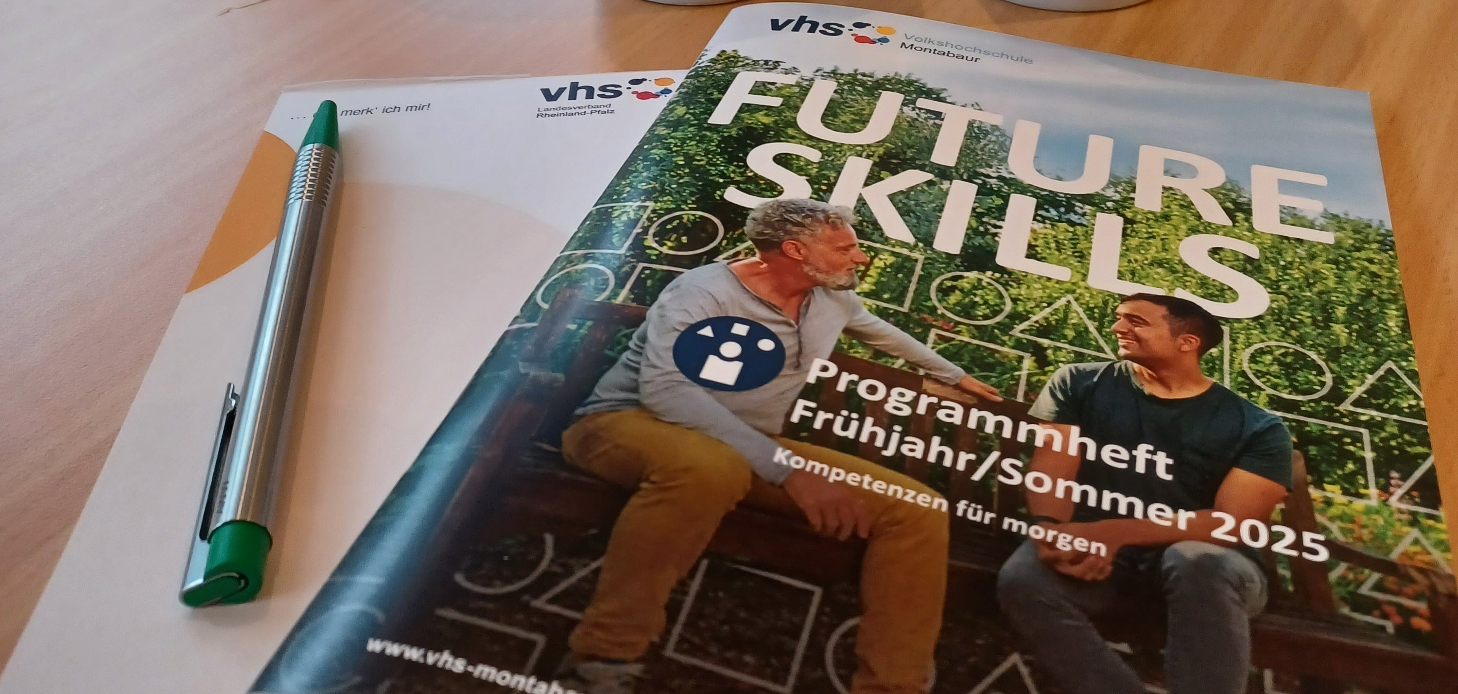 Auf einem Besprechungstisch liegen ein VHS-Programmheft mit dem Titel „FUTURE SKILLS – Programmheft Frühjahr/Sommer 2025“, ein Notizblock mit einem Stift sowie zwei VHS-Tassen mit bunten Punkten. Im Hintergrund steht eine dekorative Pflanze mit gelben Bändern. Die Szene vermittelt eine einladende und professionelle Atmosphäre, vermutlich in einer Volkshochschule oder einem Bildungszentrum.