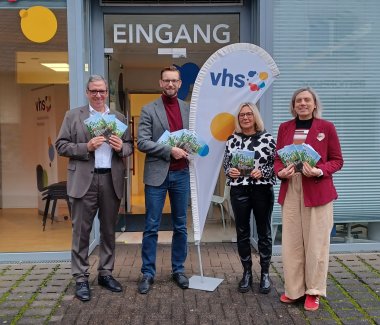 Vier Personen stehen vor dem Eingang einer Volkshochschule (VHS) und halten Flyer oder Broschüren in den Händen. Hinter ihnen ist eine Fahne mit dem VHS-Logo sowie ein Schild mit der Aufschrift „EINGANG“ zu sehen. Die Gruppe wirkt offiziell gekleidet und präsentiert das Programm für das neue Semester.