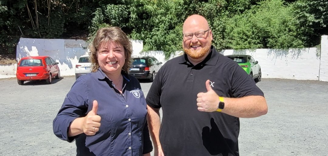 Zwei Personen stehen nebeneinander auf einem Schotterparkplatz und zeigen Daumen hoch in die Kamera. Im Hintergrund sind mehrere geparkte Autos und eine von Bäumen gesäumte Böschung zu sehen, die dichten grünen Wald zeigt. Beide Personen wirken fröhlich und der sonnige Tag sorgt für kräftige Schatten. 