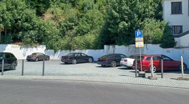 Ein kleiner Parkplatz mit mehreren geparkten Autos auf einem Schotteruntergrund, umgeben von dichter, grüner Böschung und einem weißen Mauerabschnitt. Rechts im Bild steht ein blaues Parkschild mit dem Hinweis „nur mit Parkschein“, daneben ein Wohnhaus mit Balkonen. Die Szene wirkt ruhig und sonnig. 