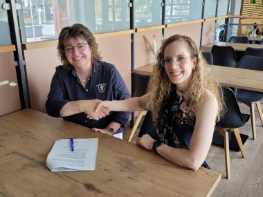 Zwei Frauen sitzen sich an einem Holztisch in einem modernen Café oder Restaurant gegenüber und schütteln sich lächelnd die Hand, vor ihnen liegt ein unterzeichneter Vertrag mit einem blauen Kugelschreiber. Die Szene wirkt freundlich und offiziell, als Abschluss einer erfolgreichen Vereinbarung. 