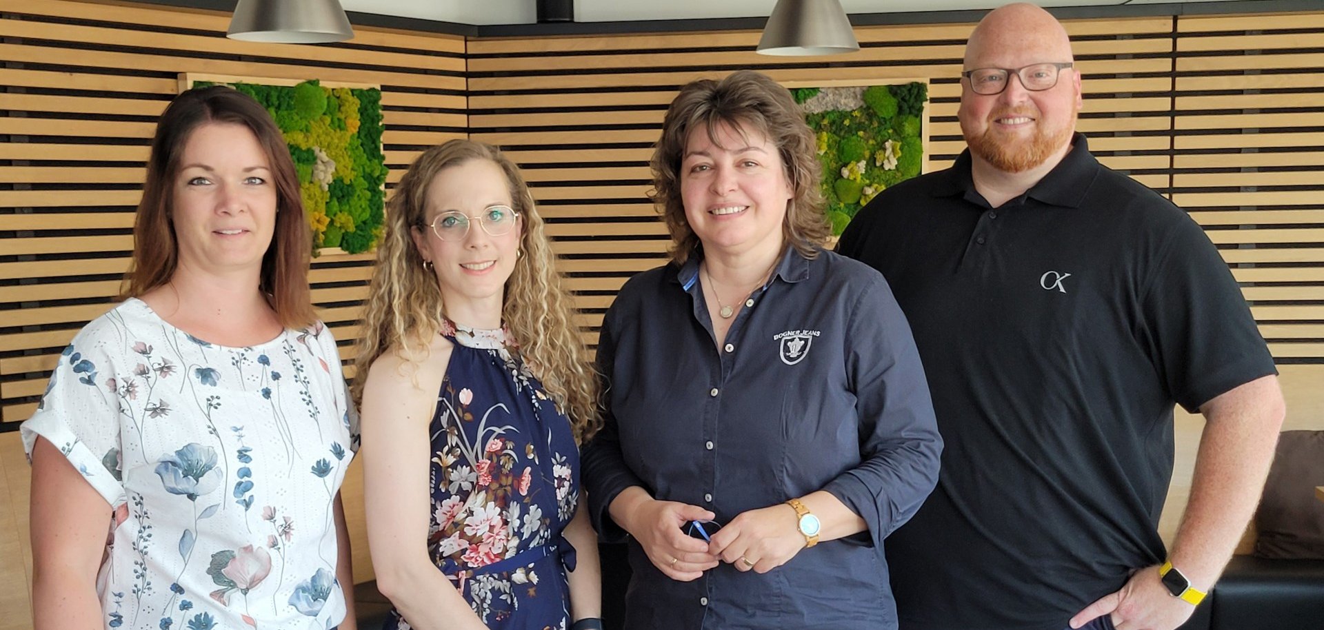 Vier Personen stehen nebeneinander in einem modern eingerichteten Raum mit Holzverkleidung und grünen Wandakzenten, alle lächeln in die Kamera. Zwei Frauen tragen geblümte Outfits, eine Frau ein dunkelblaues Hemd mit Logo, der Mann ein schwarzes Poloshirt – die Szene wirkt freundlich und professionell, vermutlich im Rahmen eines geschäftlichen Treffens. 