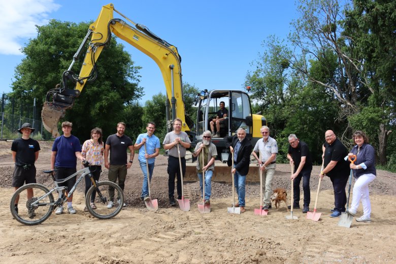 Gruppenfoto von etwa 13 Personen beim Spatenstich für ein Bauprojekt, darunter Jugendliche, Erwachsene und ein Hund, mit einem gelben Bagger im Hintergrund und einem Mountainbike im Vordergrund. Einige Beteiligte halten Spaten in den Händen und lächeln, während ein Mann im Bagger sitzt – symbolischer Startschuss für den Bau einer Pumptrack-Anlage.