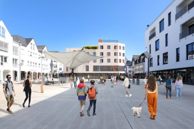 Visualisierung eines modernen Stadtplatzes in Montabaur mit der geplanten City-Jugendherberge im Hintergrund, einem runden, mehrstöckigen Gebäude mit schlichten Fenstern und einem begrünten Dach. Menschen flanieren über den Platz, darunter Kinder, Familien und eine Frau mit einem kleinen weißen Hund, während im Zentrum ein großes, schirmartiges Sonnensegel Schatten spendet. 