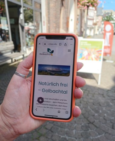 Eine Hand hält ein Smartphone, auf dem die Website gelbachtal.info mit dem Slogan „Natürlich frei – Gelbachtal“ angezeigt wird.