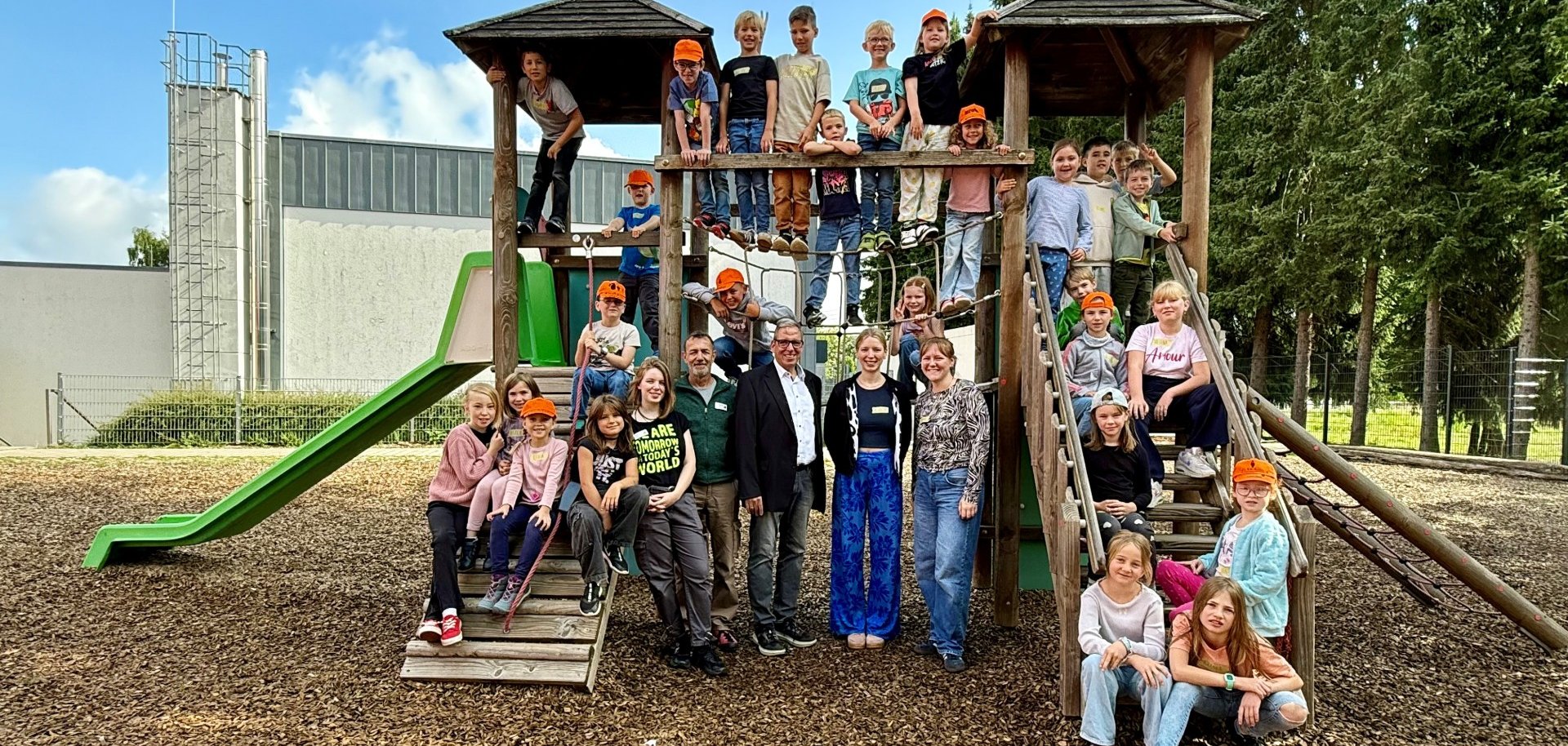 Eine große Gruppe von Kindern und Erwachsenen posiert fröhlich auf und um ein hölzernes Klettergerüst mit zwei Türmen und Rutsche auf einem Spielplatz. Viele Kinder tragen orangefarbene Kappen, einige sitzen auf der Rutsche oder hängen an Seilen, während die Erwachsenen davor stehen und lachen. Im Hintergrund sind Bäume und ein Gebäude mit grauer Fassade zu sehen.