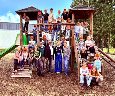 Eine große Gruppe von Kindern und Erwachsenen posiert fröhlich auf und um ein hölzernes Klettergerüst mit zwei Türmen und Rutsche auf einem Spielplatz. Viele Kinder tragen orangefarbene Kappen, einige sitzen auf der Rutsche oder hängen an Seilen, während die Erwachsenen davor stehen und lachen. Im Hintergrund sind Bäume und ein Gebäude mit grauer Fassade zu sehen.