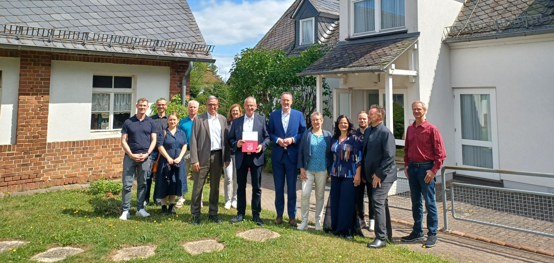 Innenminister Ebling übergibt in Gackenbach einen Förderbescheid an Ortsbürgermeister Weidenfeller. Gruppenfoto vor Wohnhäusern.