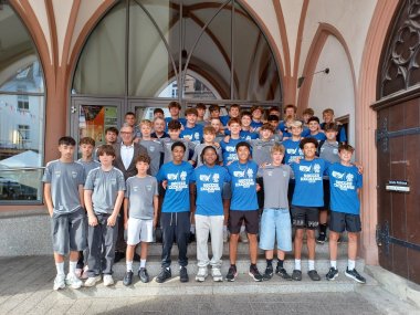 Eine große Gruppe Jugendlicher, hauptsächlich Jungen, steht auf einer Steintreppe vor einem historischen Gebäude mit gotischen Bögen. Viele tragen blaue T-Shirts mit der Aufschrift „Soccer Exchange“, andere graue Trikots mit Vereinslogo. In der ersten Reihe steht auch ein Mann im Anzug, vermutlich ein offizieller Vertreter oder Gast des Fußballaustauschs.