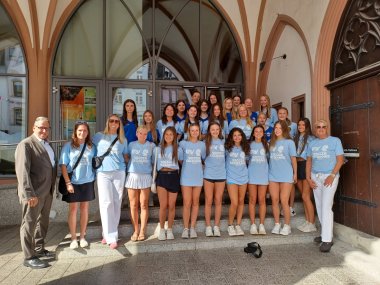 Eine Gruppe junger Frauen steht gemeinsam mit mehreren Erwachsenen auf einer Treppe vor einem historischen Gebäude mit gotischen Bögen. Die meisten Teilnehmerinnen tragen hellblaue T-Shirts mit der Aufschrift „Soccer Exchange“, was auf ein internationales Fußballaustauschprogramm hinweist. Zwei Männer in Anzügen sowie weitere Betreuerinnen in Sportkleidung flankieren die Gruppe.
