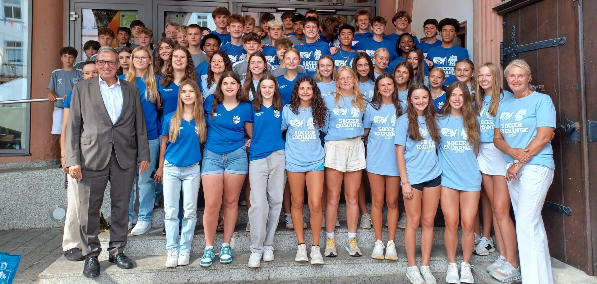Eine große Gruppe Jugendlicher und Erwachsener posiert auf einer Treppe vor einem historischen Gebäude mit gotischen Bögen. Die Jugendlichen tragen überwiegend blaue T-Shirts mit der Aufschrift „Soccer Exchange“, was auf ein Austauschprogramm im Fußball hindeutet. Links und rechts stehen zwei Erwachsene, einer im Anzug und die andere in weißer Hose und ebenfalls „Soccer Exchange“-Shirt.