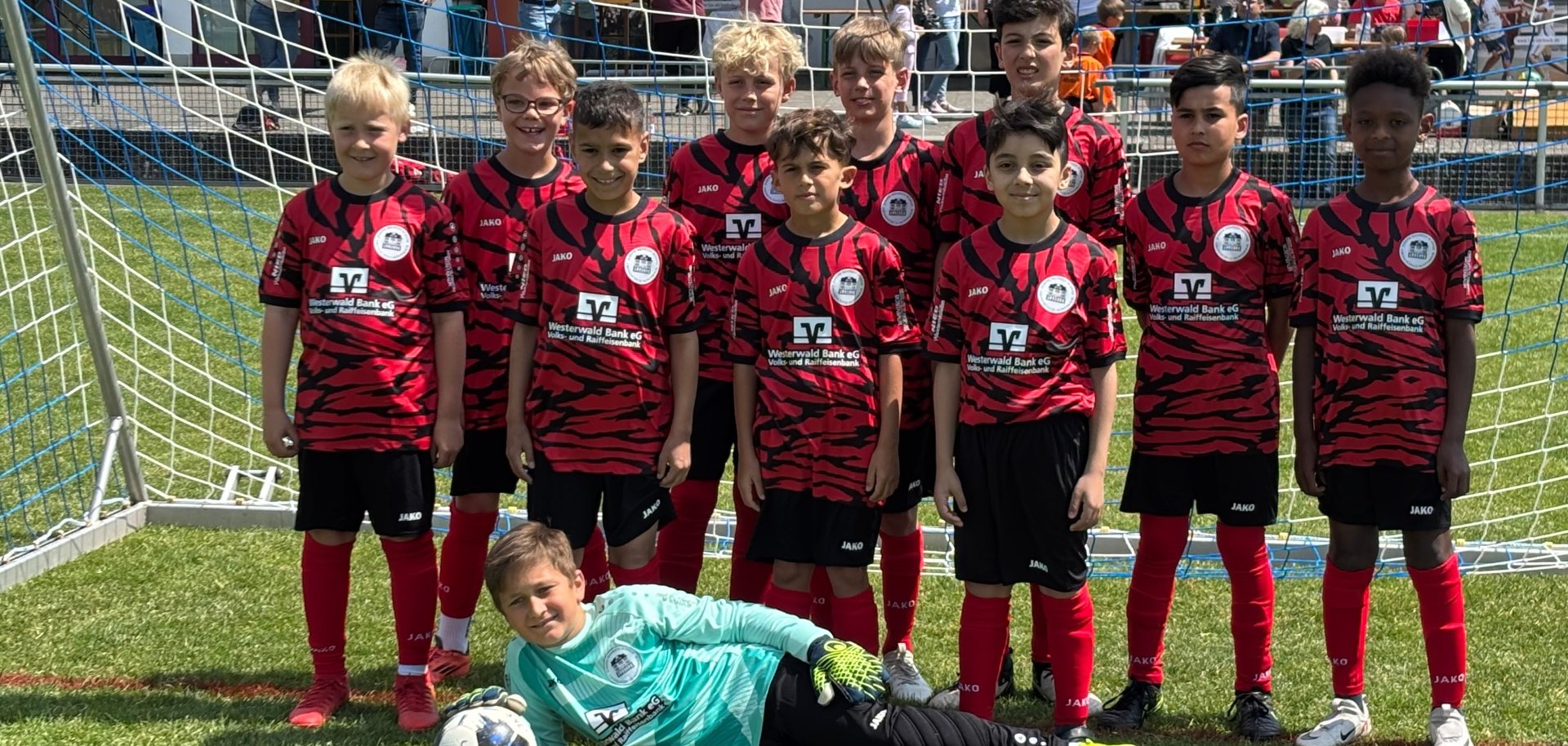 Eine Sieger-Jugendfußballmannschaft in rot-schwarzen Trikots steht vor einem Tor, während ein Junge im Torwarttrikot mit einem Ball davor liegt. Im Hintergrund sind Zuschauer zu sehen. 