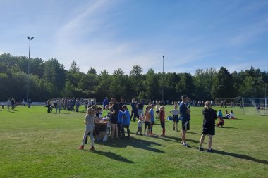 Viele Kinder und Erwachsene versammeln sich auf einem Sportplatz. Kinder in bunten Sportklamotten spielen und unterhalten sich. Im Hintergrund sind weitere Zuschauergruppen und Bäume zu sehen.