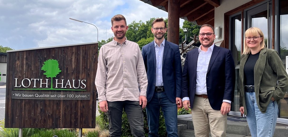 Vier Personen stehen vor dem Firmensitz von Loth Haus neben einem Schild mit der Aufschrift „Loth Haus – Wir bauen Qualität seit über 100 Jahren“.