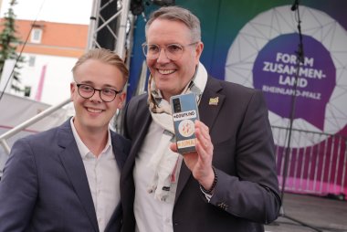 Maxim Chetchouga und Ministerpräsident Alexander Schweitzer, der ein Handy mit Jumazu-Aufkleber in die Kamera hält.