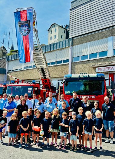 Gruppenfoto vor dem Feuerwehrhaus Montabaur mit mehreren Feuerwehrfahrzeugen im Hintergrund. Vor der Drehleiter mit großer „Freiwillige Feuerwehr Montabaur“-Fahne stehen zahlreiche Kinder in dunklen T-Shirts, begleitet von Feuerwehrleuten und Betreuenden – vermutlich zur Gründung einer Bambini-Feuerwehr. Im Hintergrund sind auch Altbauten und ein blauer Himmel zu sehen.