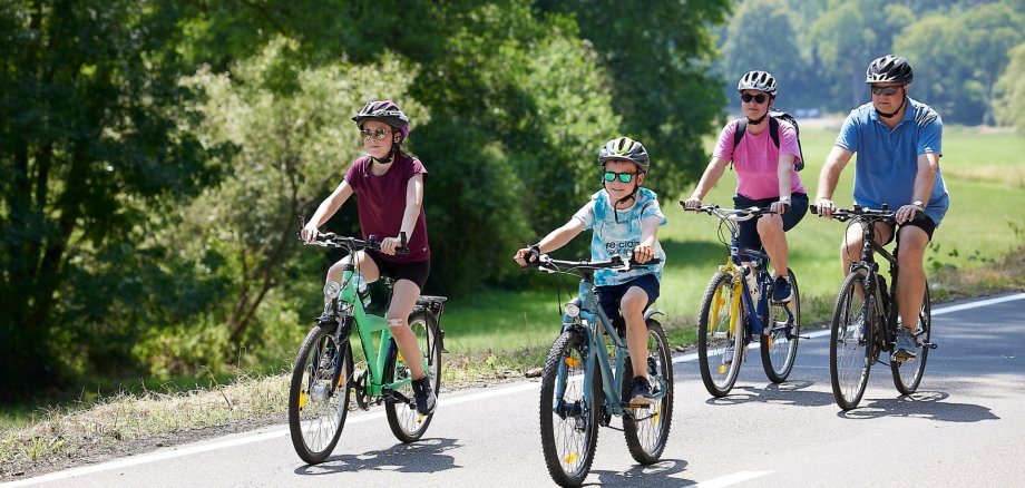 Vier Personen, zwei Kinder und zwei Erwachsene, fahren bei sonnigem Wetter gemeinsam Fahrrad auf einer Landstraße durch eine grüne, ländliche Landschaft. Alle tragen Fahrradhelme und Sonnenbrillen und wirken entspannt und fröhlich. 