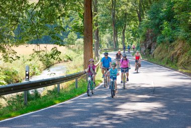 Mehrere Menschen fahren gemeinsam Fahrrad auf einer von Bäumen gesäumten Landstraße entlang eines kleinen Bachs. Die Szene wirkt sommerlich und entspannt, mit Erwachsenen und Kindern, die Helme tragen und das schöne Wetter genießen. 
