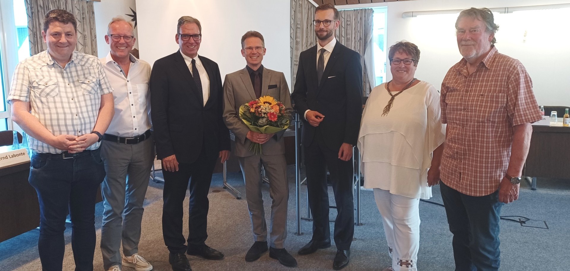 Sieben Personen stehen nebeneinander in einem Besprechungsraum und lächeln in die Kamera. Der Mann in der Mitte hält einen bunten Blumenstrauß mit einer Sonnenblume – er wurde offenbar soeben geehrt oder gewählt. Die Gruppe ist gemischt gekleidet, von festlich bis leger, und im Hintergrund sind Tische, Namensschilder und eine Leinwand sichtbar. 