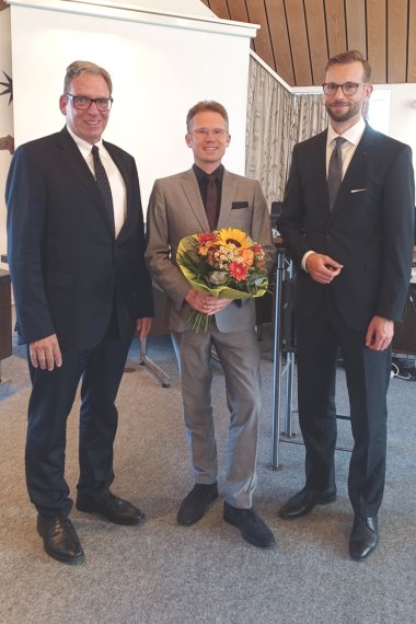 Drei Männer in Anzügen stehen in einem Besprechungsraum und lächeln in die Kamera. Der Mann in der Mitte hält einen farbenfrohen Blumenstrauß mit Sonnenblume, Rosen und Schleierkraut – vermutlich wurde er gerade geehrt oder gewählt. Im Hintergrund ist eine Leinwand sowie technische Ausstattung zu sehen. 
