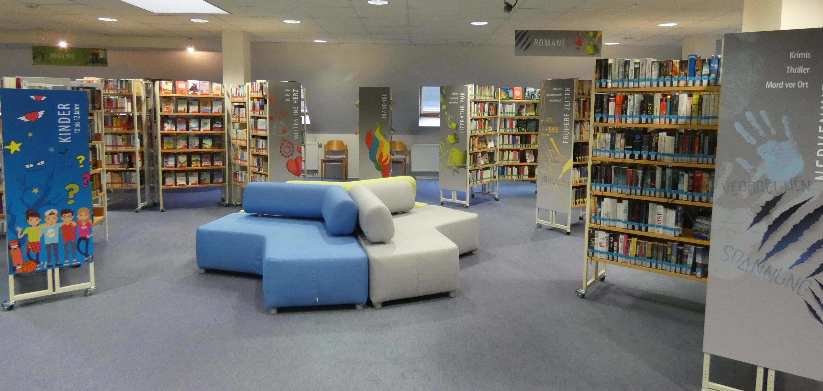 Die Aufnahme zeigt den Innenraum einer Bibliothek mit Regalen voller Bücher und einer modernen Sitzecke in Blau und Grau in der Mitte. Verschiedene Bereichsschilder wie „Kinder“, „Romane“ und „Jugend“ weisen auf die thematische Aufteilung der Büchersammlungen hin. Die Bibliothek ist hell beleuchtet und wirkt einladend für Leserinnen und Leser aller Altersgruppen.