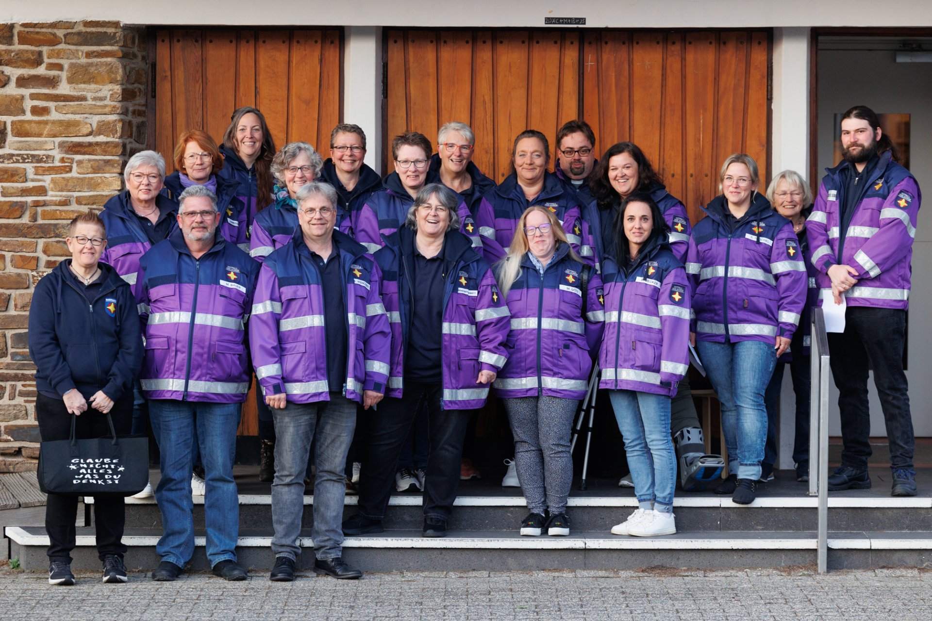 Gruppenfoto von etwa zwanzig Personen, die größtenteils eine einheitliche, violett-blaue Jacke mit reflektierenden Streifen tragen – vermutlich Notfallseelsorger*innen oder ein Hilfsteam. Sie stehen auf einer Treppe vor einem Gebäude mit Holz- und Steinfassade, lächeln in die Kamera. Eine Person links hält eine schwarze Tasche mit weißer Aufschrift: „Glaube nicht alles, was du denkst.“