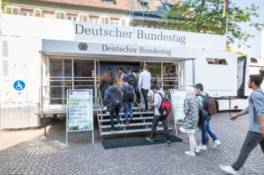 Eine Gruppe junger Menschen betritt einen mobilen Infostand des Deutschen Bundestags, der in einem weißen Truck mit Rampe und Treppe untergebracht ist. Zwei Aufsteller neben dem Eingang zeigen Öffnungszeiten und Hinweise zur Ausstellung über das deutsche Parlament.