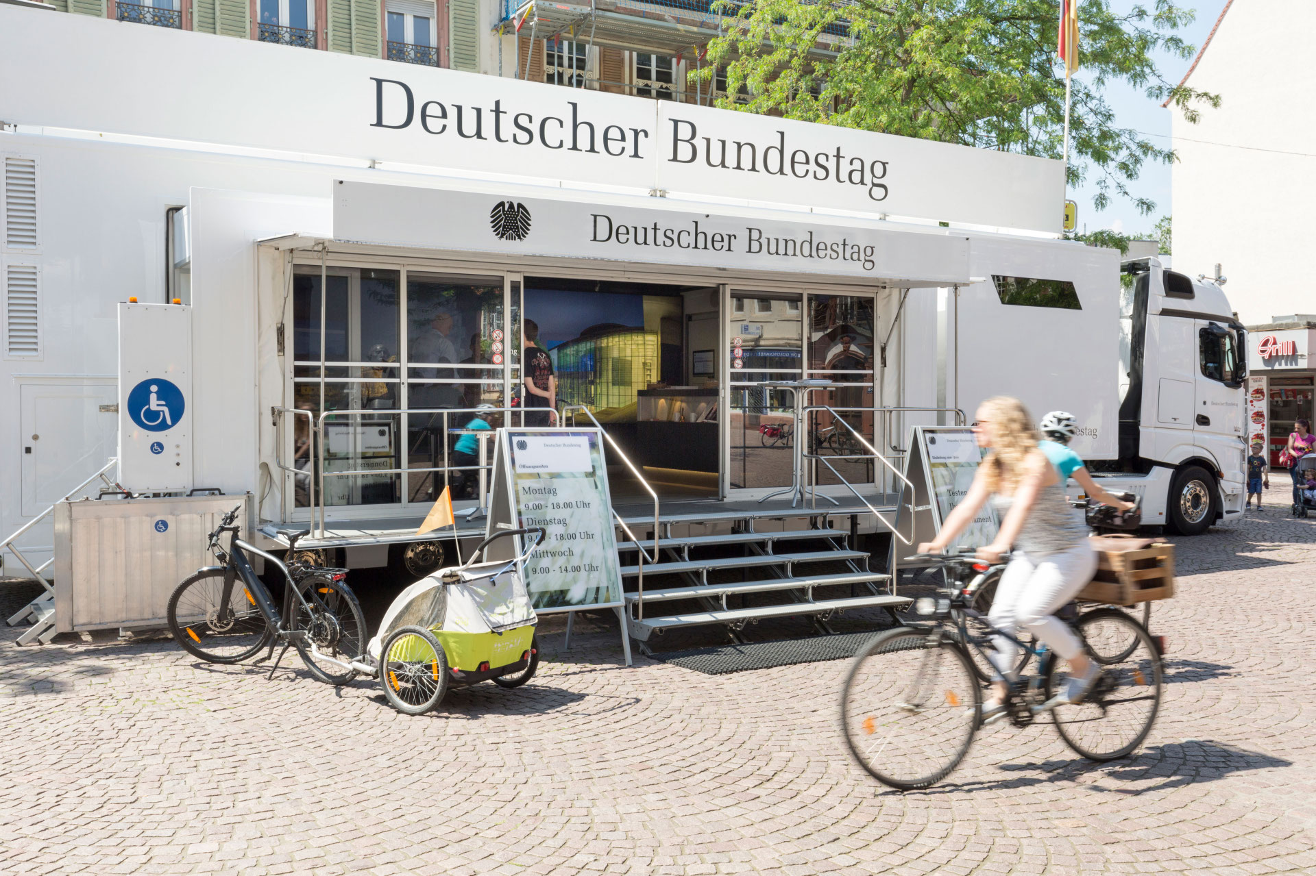 Ein mobiler Infostand des Deutschen Bundestags in einem weißen Truck ist auf einem Platz aufgebaut, mit zwei Aufstellern, die Öffnungszeiten anzeigen. Vor dem Eingang fahren zwei Personen auf Fahrrädern vorbei, im Vordergrund ein Fahrrad mit einem Kindersitzanhänger.