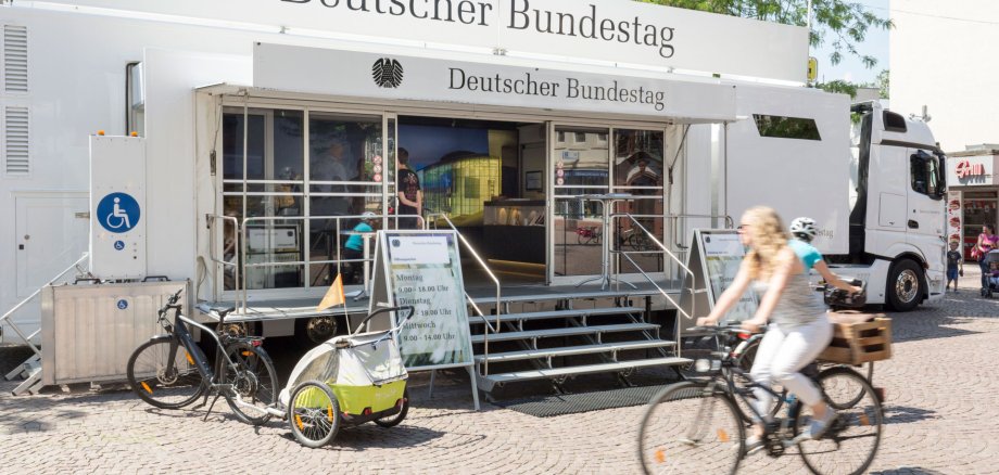 Ein mobiler Infostand des Deutschen Bundestags in einem weißen Truck ist auf einem Platz aufgebaut, mit zwei Aufstellern, die Öffnungszeiten anzeigen. Vor dem Eingang fahren zwei Personen auf Fahrrädern vorbei, im Vordergrund ein Fahrrad mit einem Kindersitzanhänger.