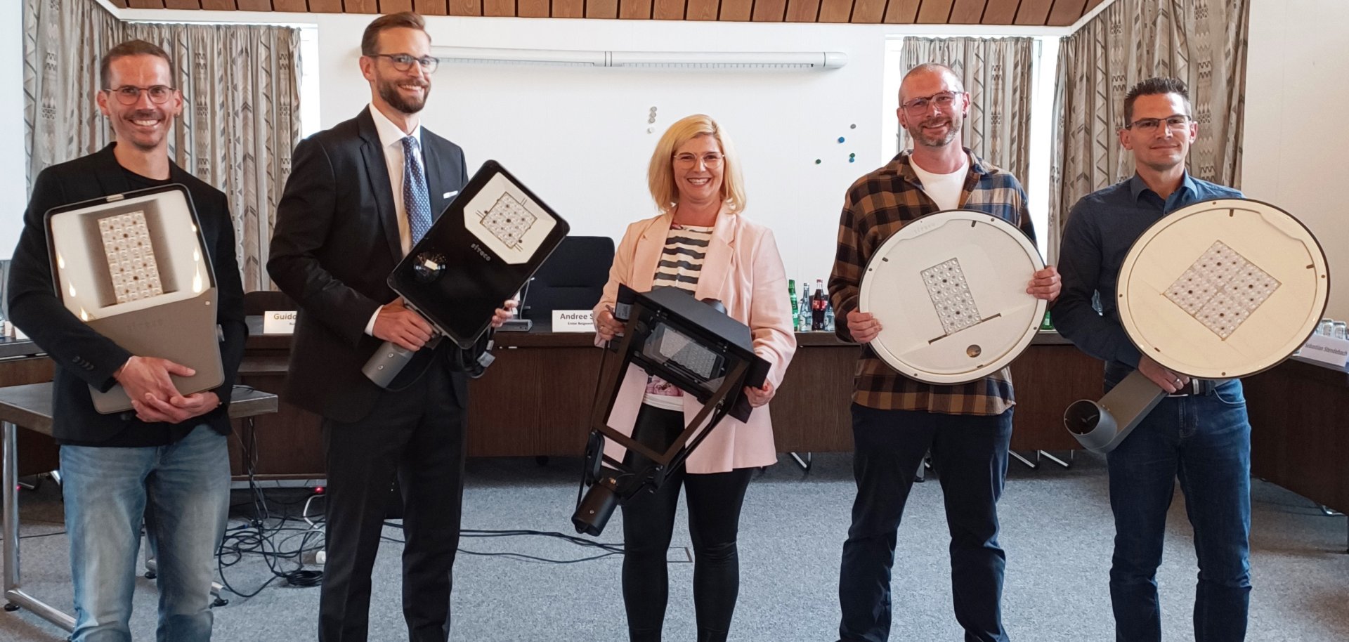 Fünf Personen stehen in einem Konferenzraum nebeneinander und halten verschiedene Modelle moderner LED-Straßenlaternen in den Händen. Die Gruppe lächelt in die Kamera, im Hintergrund sind Tische mit Namensschildern und Getränken zu sehen.